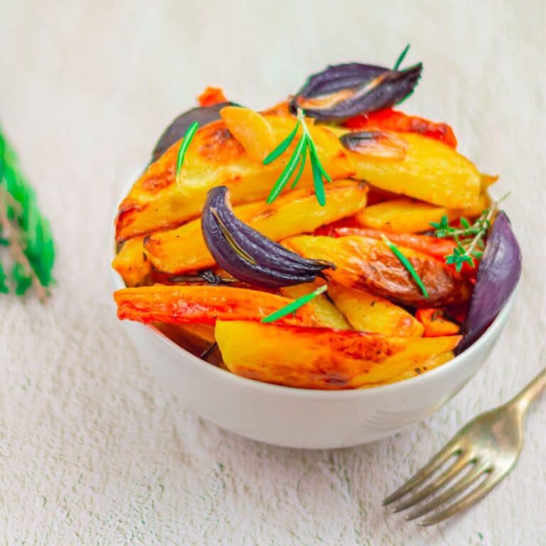 garlic herb roasted root vegetables in a white bowl placed on a tile along with herbs and a fork.