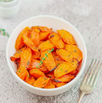 maple glazed carrots in a white bowl with a sprig of thyme placed on a ceramic along with a fork and couple of pinchbowls.