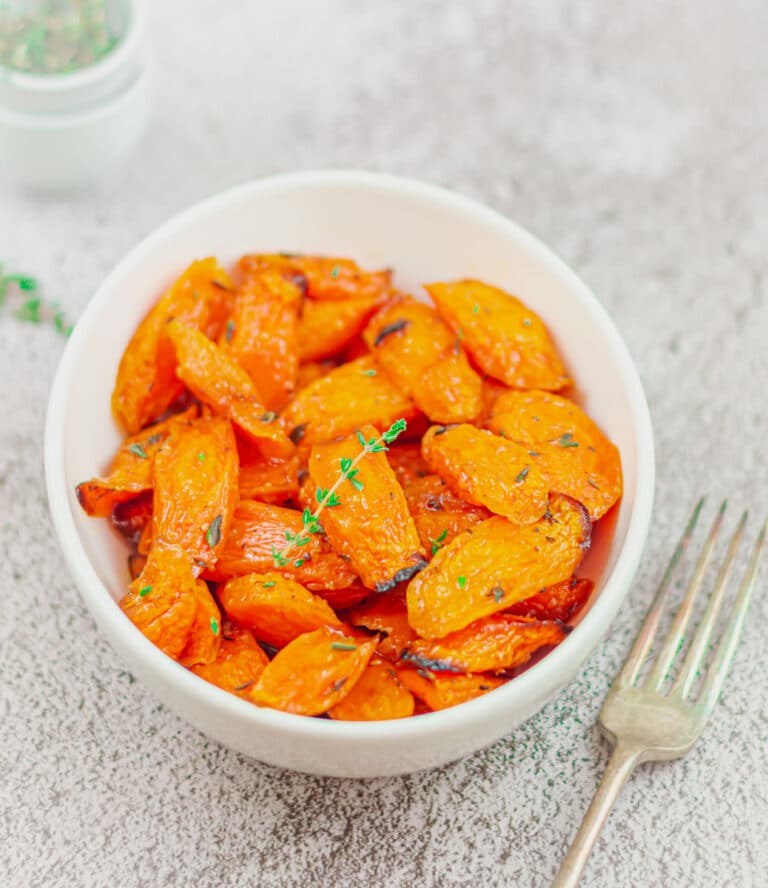 maple glazed carrots in a white bowl with a sprig of thyme placed on a ceramic along with a fork and couple of pinchbowls.