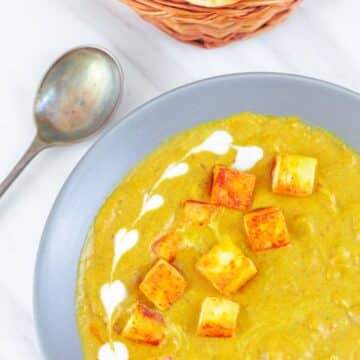 paneer korma in a grey bowl garnished with cream placed on a table along with a spoon, basket of naan breads and a cloth.