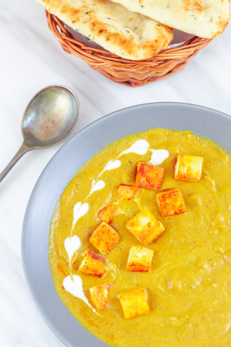 paneer korma in a grey bowl garnished with cream placed on a table along with a spoon, basket of naan breads.