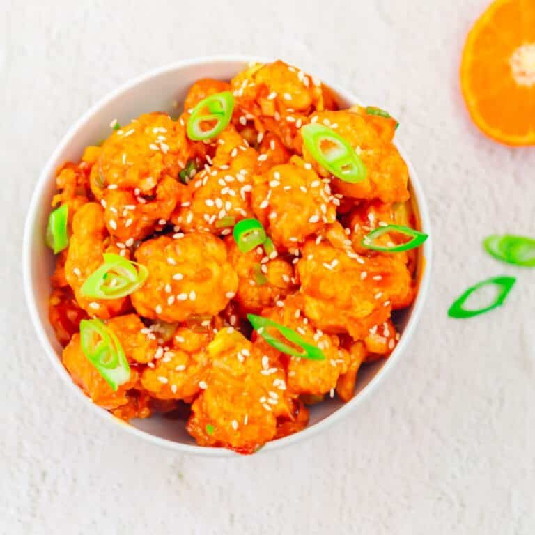 orange cauliflower in a white bowl placed on a white table with a sliced orange on side.