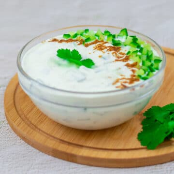 cucumber raita in a glass bowl topped with cucumber, cumin powder and coriander placed on a wooden board.