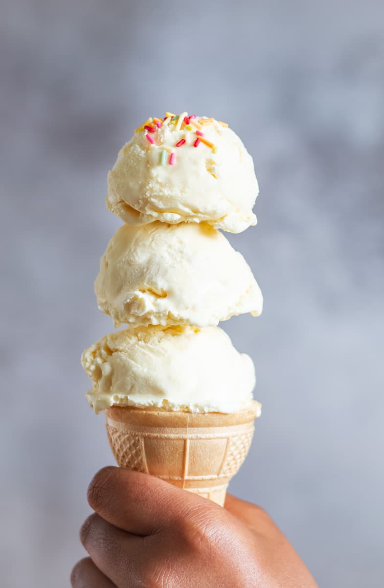 Holding a cone with 3 scoops of vanilla icecream with sprinkles on top. 
