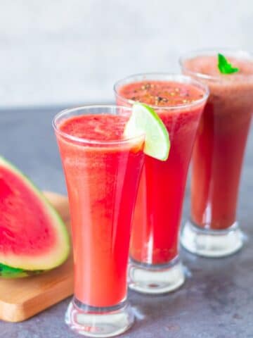 close up shot of 3 glasses watermelon juice placed on a granite along with a quartered watermelon on a wooden board.