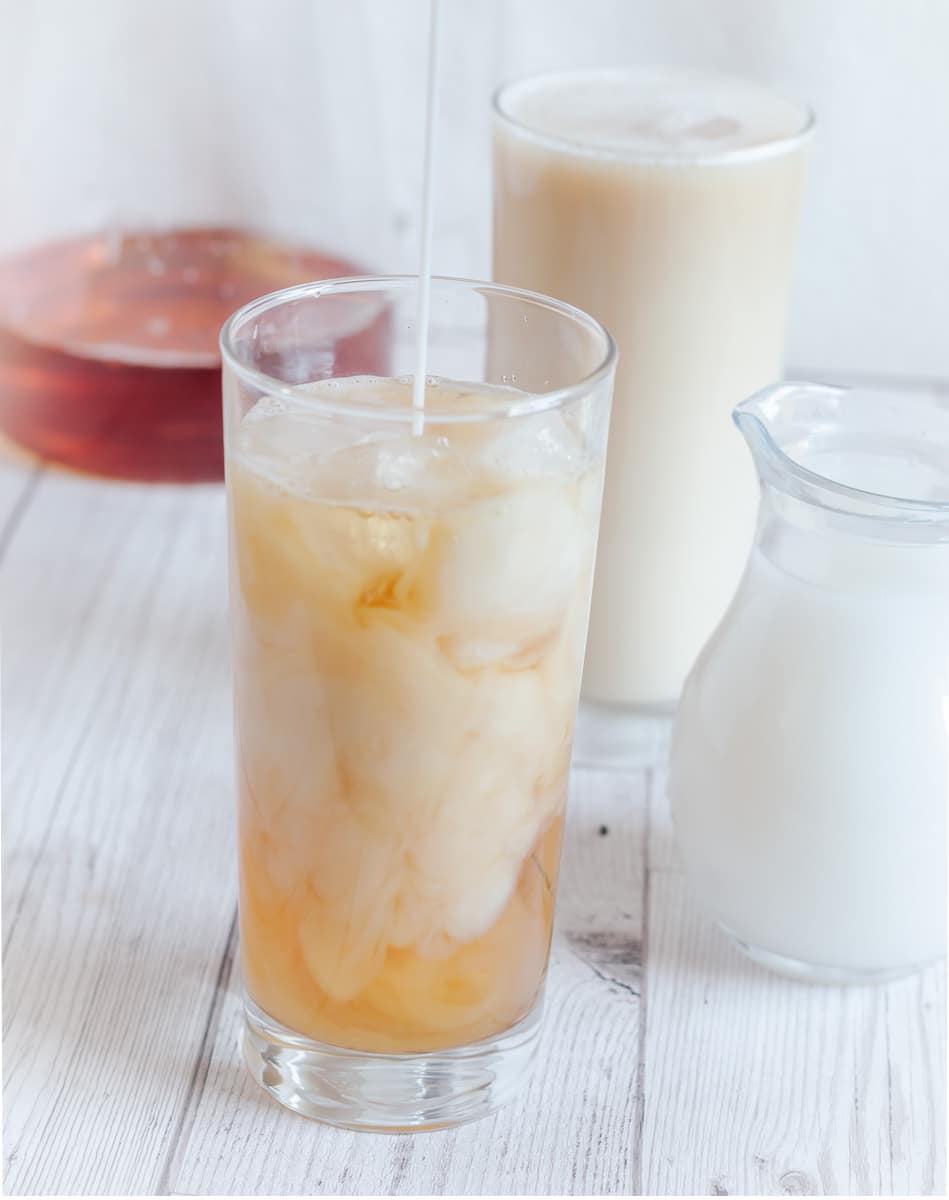 2 high ball glasses of iced london fog tea placed on a tile along with a jug of milk and a jug of black tea. 