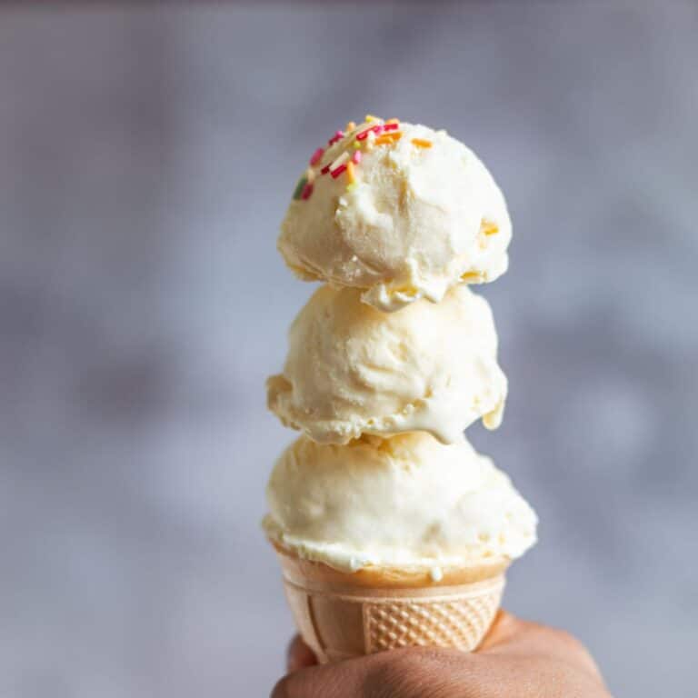 Holding a cone with 3 scoops of vanilla icecream with sprinkles on top.