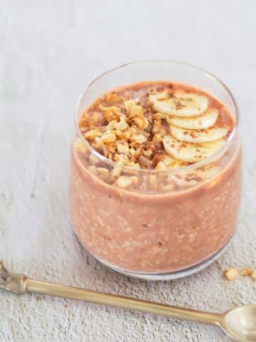 close up shot of banana chocolate overnight oats in a glass with toppings of banana slices, walnuts placed on a tile along with a spoon.