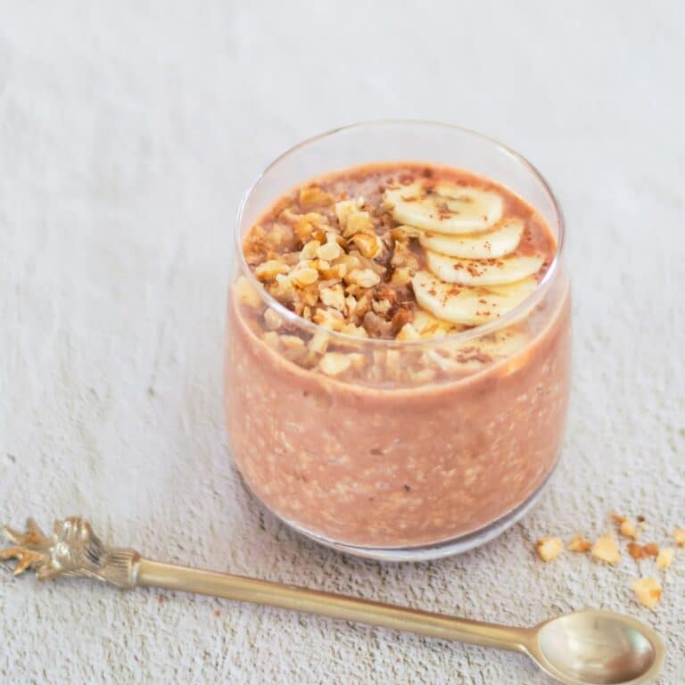 close up shot of banana chocolate overnight oats in a glass with toppings of banana slices, walnuts placed on a tile along with a spoon.