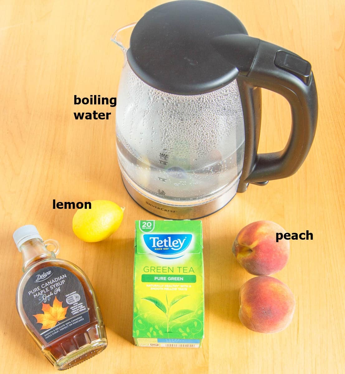 peaches, maple syrup, green tea bags, lemon and a kettle placed on a wooden table.