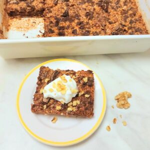 three stacked chocolate baked oats in a white plate placed on a table along with a bottle of milk and tray of baked oats.