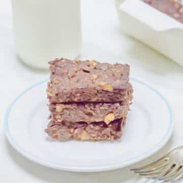 three stacked chocolate baked oats in a white plate placed on a table along with a bottle of milk and tray of baked oats.