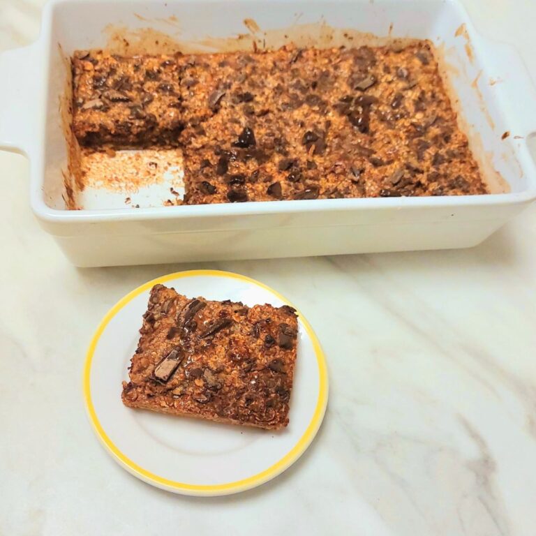 chocolate baked oats stacked in a plate placed on a table along with milk bottle and a white baking tray of baked oats.