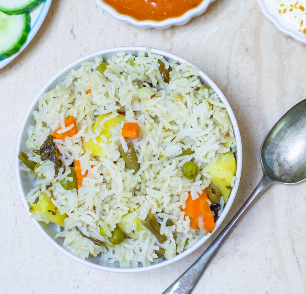 vegetable pulao in a white bowl placed on a tile along with a spoon, salad, curry and raita.