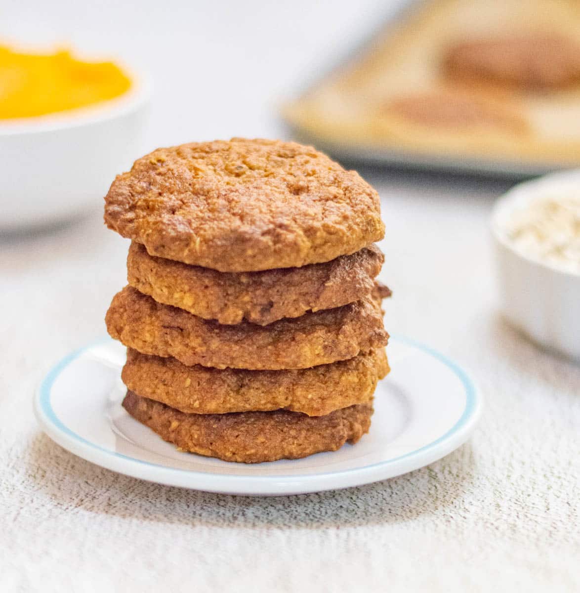 stack of pumpkin oatmeal cookies in a white plate placed on a table along with pumpkin puree, oats and a tray of cookies.