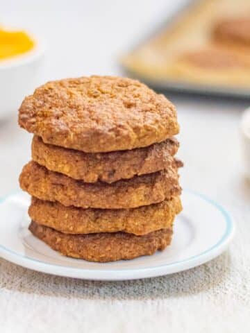 stack of 5 pumpkin oatmeal cookies in a white plate placed on a table along with pumpkin puree, oats and a tray of cookies.