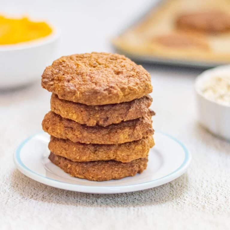 stack of 5 pumpkin oatmeal cookies in a white plate placed on a table along with pumpkin puree, oats and a tray of cookies.