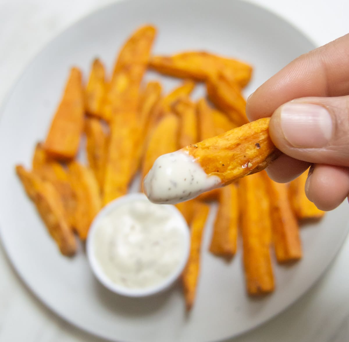 a hand holding a sweet potato wedge dipped in a white sauce on top of a plate of sweet potato wedges.