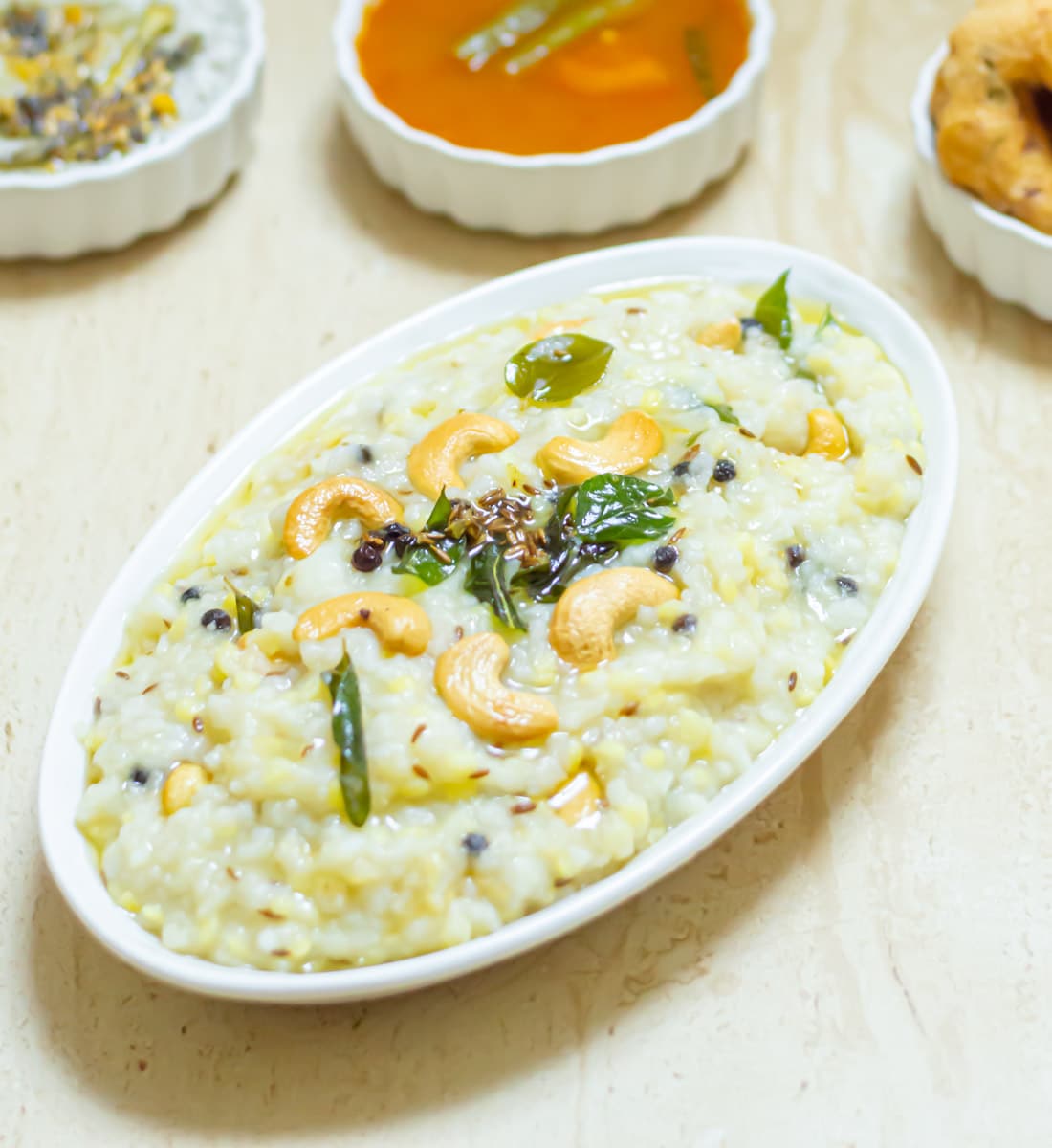 ven pongal in an oval bowl placed on a tile along with a bowls of chutney, sambar and vada.