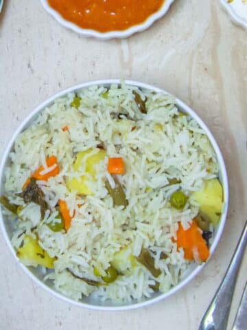 long shot of vegetable pulao in a white bowl placed on a tile along with a spoon, cucumber, curry and raita.