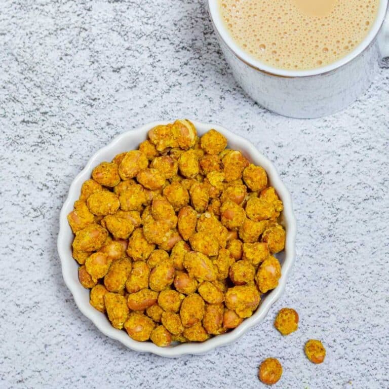 Top shot of masala peanuts in a white bowl placed on a tile along with a cup of tea.