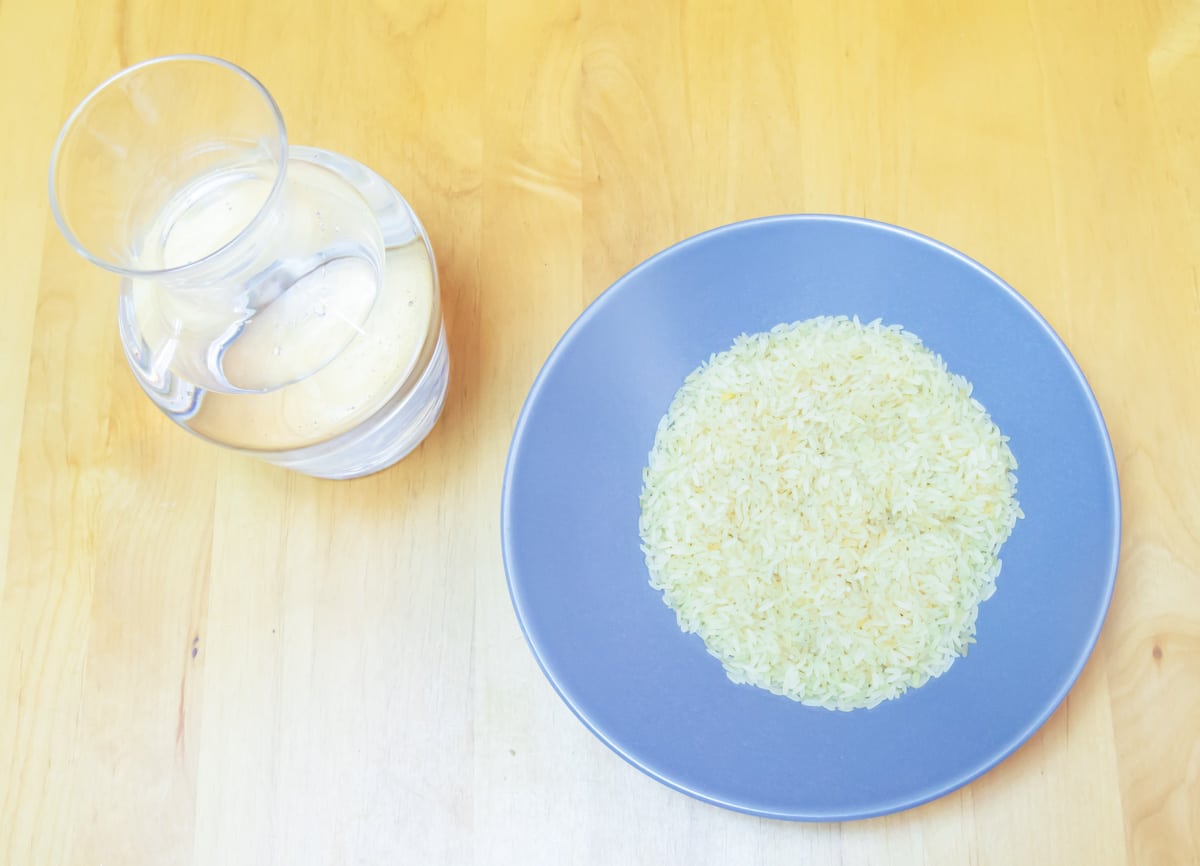 a blue bowl of raw rice and a glass jar of water placed on a wooden table.