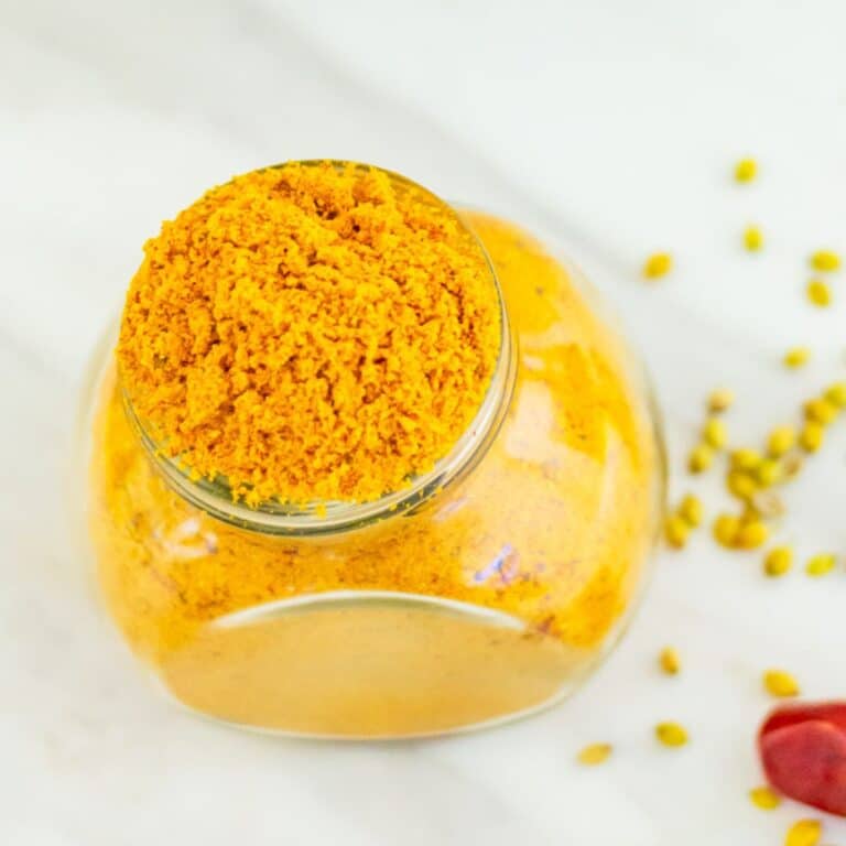 top shot of sambar masala in a glass jar placed on a marble along with a red chilli and coriander seeds next to it.