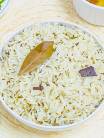 close up shot of jeera rice in a white bowl placed on a tile along with a plate of cucumber slices and a bowl of curry.