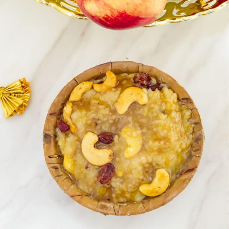top shot of sweet pongal in a sal leaf bowl placed on a marble along with a fruit basket behind.