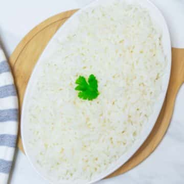 oval dish of rice with a coriander leaf placed on a wooden board along with a napkin on a marble.