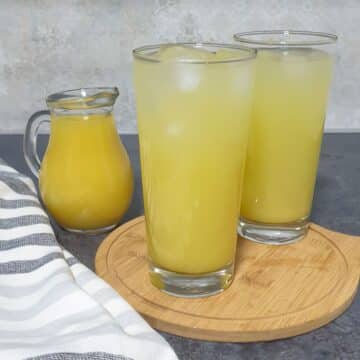 2 glasses of pineapple ginger juice placed on a wooden board with a jug of syrup behind it.