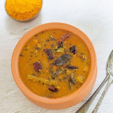 sambar in a brown pot place don awhite tile along with 2 spoons and a bowl of spice powder.