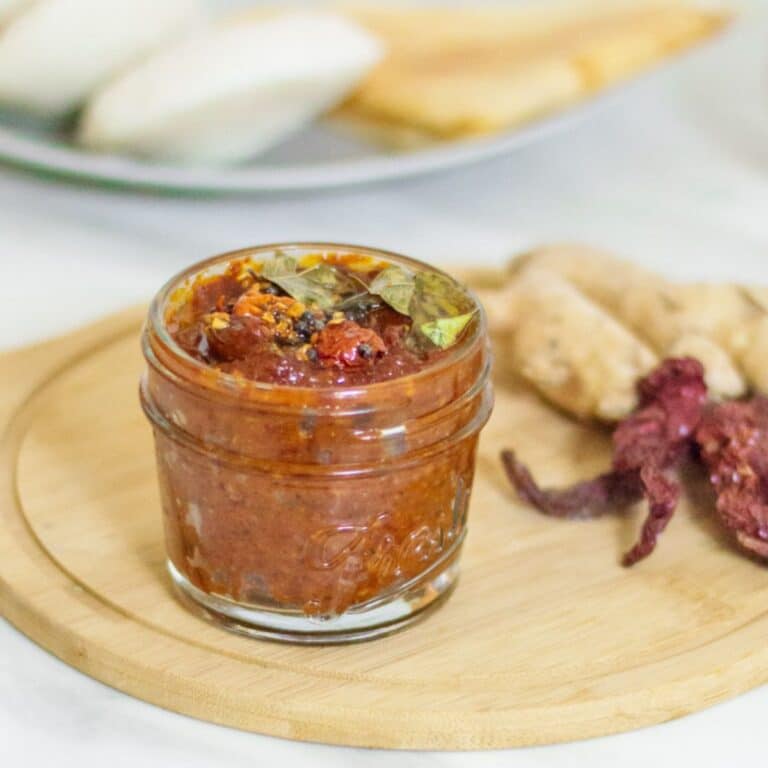 a small bottle of ginger chutney placed on a wooden board along with a few dried chillies and ginger root next to it and a plate of dosa and idlis behind it.