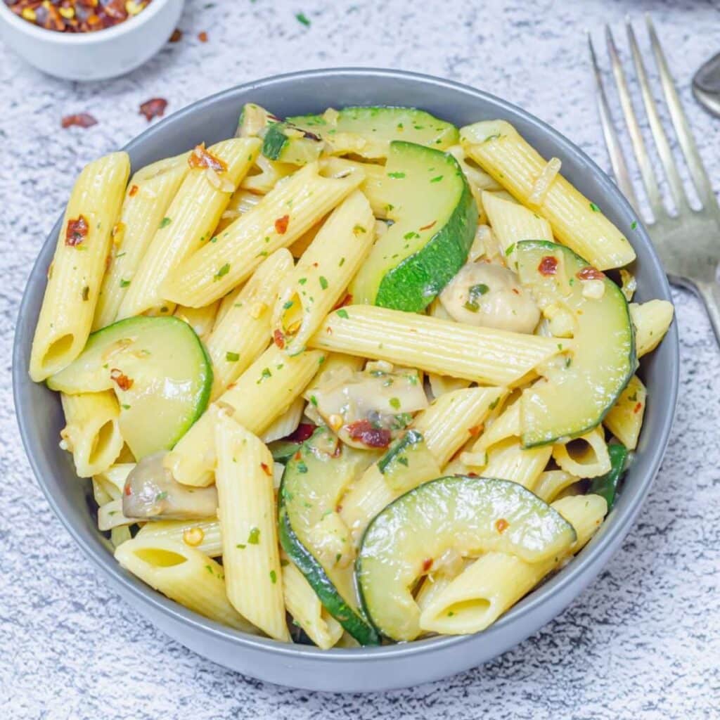 zucchini mushroom pasta in a grey bowl placed on a tile along with forks and a pinch bowl of chilli flakes.