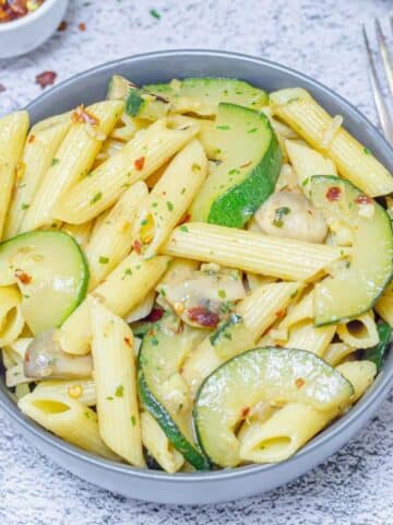 zucchini mushroom pasta in a grey bowl placed on a tile along with forks and a pinch bowl of chilli flakes.
