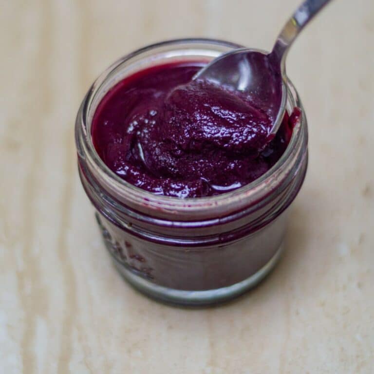 scooping out blue fruit sauce with a spoon from the glass jar placed on the tile.