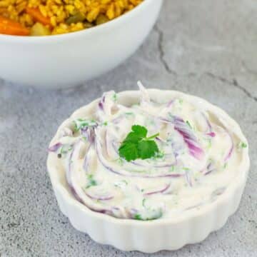 A white bowl of onion raita topped with a coriander leaf placed on a tile along with a bowl of biryani..
