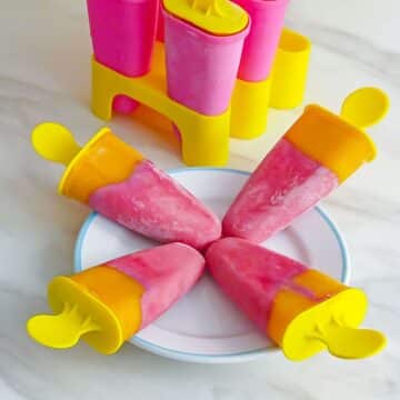four pink and yellow popsicles on a white plate placed on a marble along with popsicle base behind them.