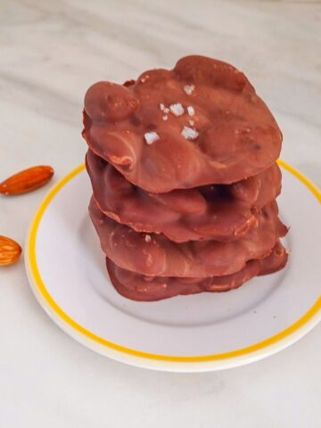 stack of chocolate almond clusters in a plate placed on a marble with a few almonds next to it.