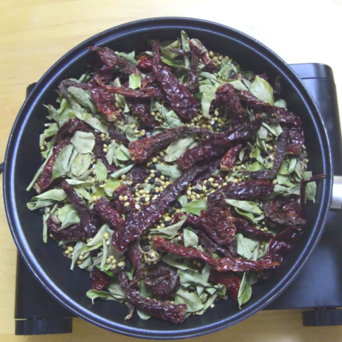 curry leaves, red chillies and whole spices in a black pan placed on a burner.
