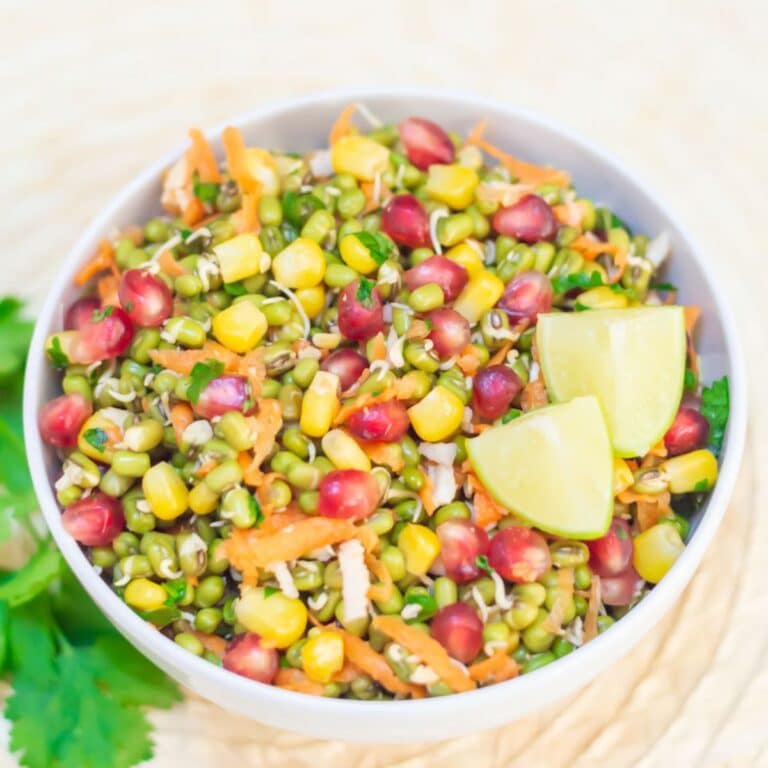 close up shot of white bowl of moong sprouts salad with 2 lemon wedges placed on a brown tile with a coriander sprig.