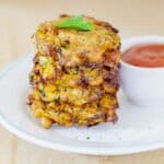 zucchini corn fritters stacked in a white plate placed on the granite along with a bowl of ketchup.