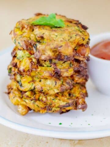 zucchini corn fritters stacked in a white plate placed on the granite along with a bowl of ketchup.