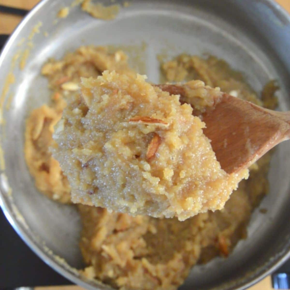 holding a wooden spoon of badam halwa on the top of steel pan of halwa.