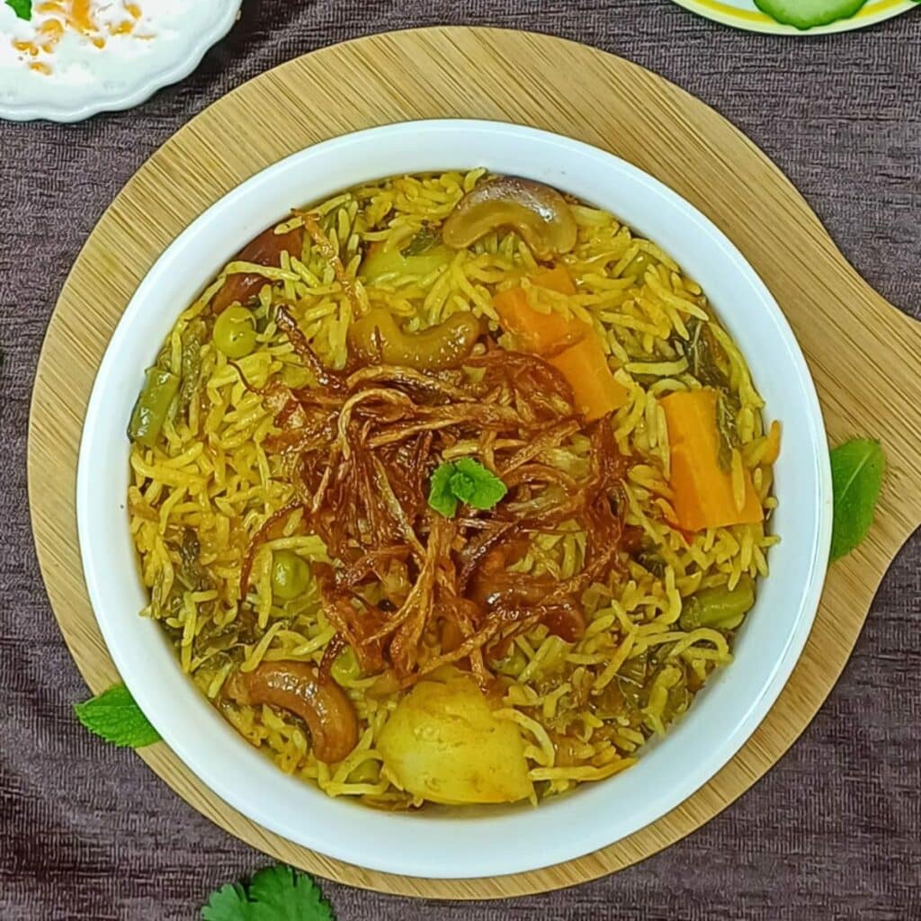 white pot of veggie biryani placed on the wooden board along with a bowl of yogurt and a plate of cucumber slices.