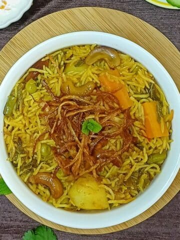 white pot of veggie biryani placed on the wooden board along with a bowl of yogurt and a plate of cucumber slices.