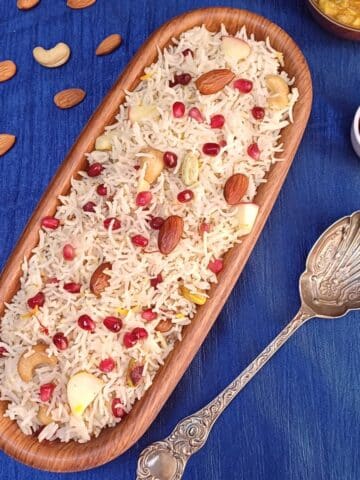 oval wooden bowl of kashmiri pulao placed on a blue cloth along with a spoon, pinch bowl of pomegranate and few nuts.