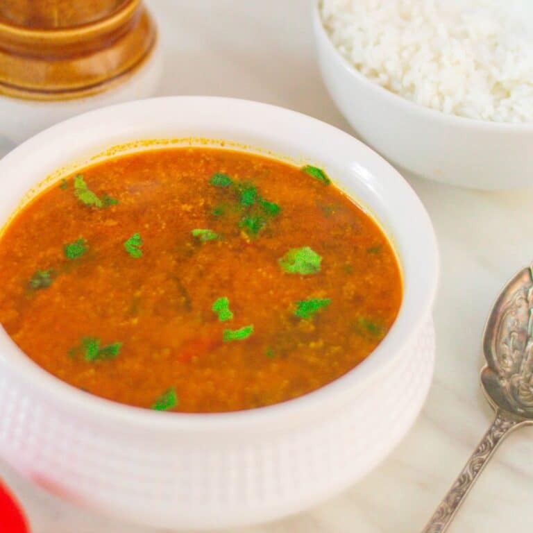 white bowl of tomato rasam placed on marble along with a spoon, rice bowl, and pickle bowl.