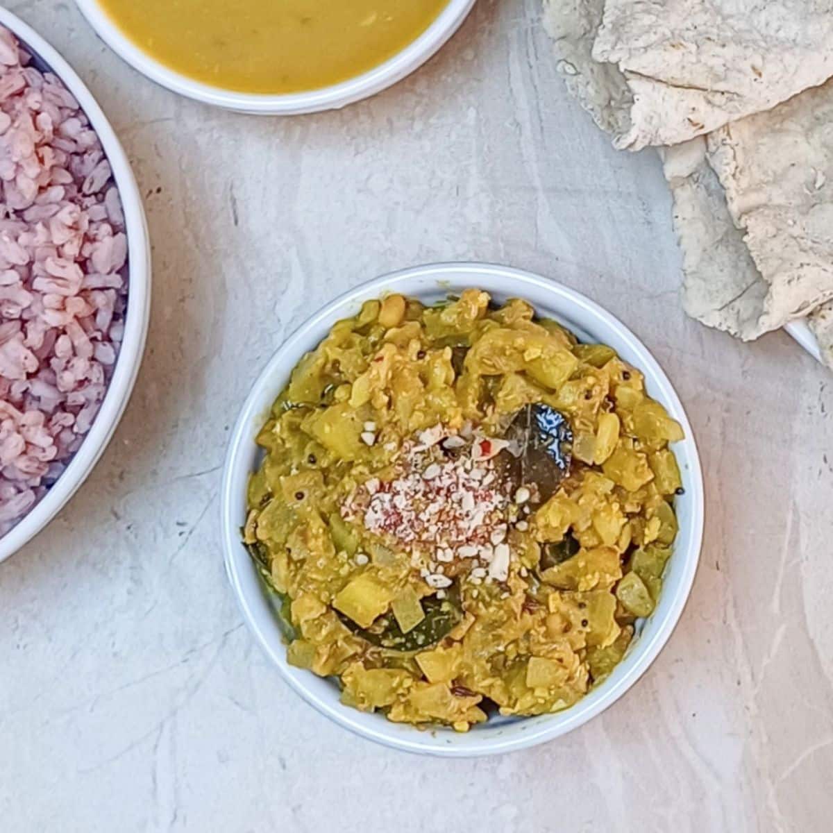 lauki ki sabji in a white bowl placed on a tile along with a bowl of red rice, curry and rotis.