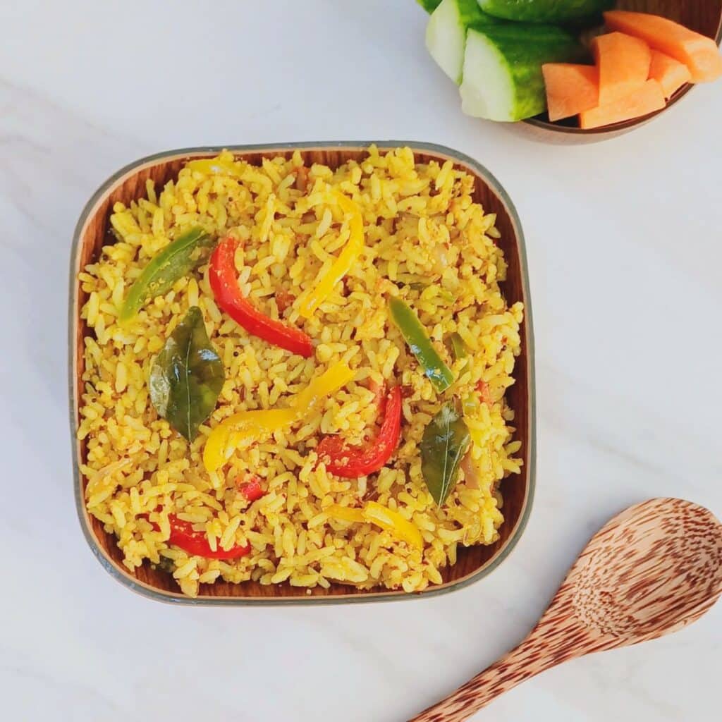 square bowl filled with capsicum rice placed on marble along with a wooden spoon and bowl of vegetable sticks.